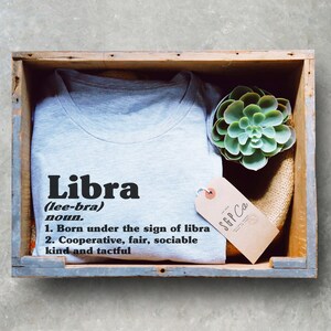 May include: A gray t-shirt with the word "Libra" in black text and a definition of the word. The t-shirt is in a wooden box with a small succulent plant and a brown tag with the text "SGP Co."