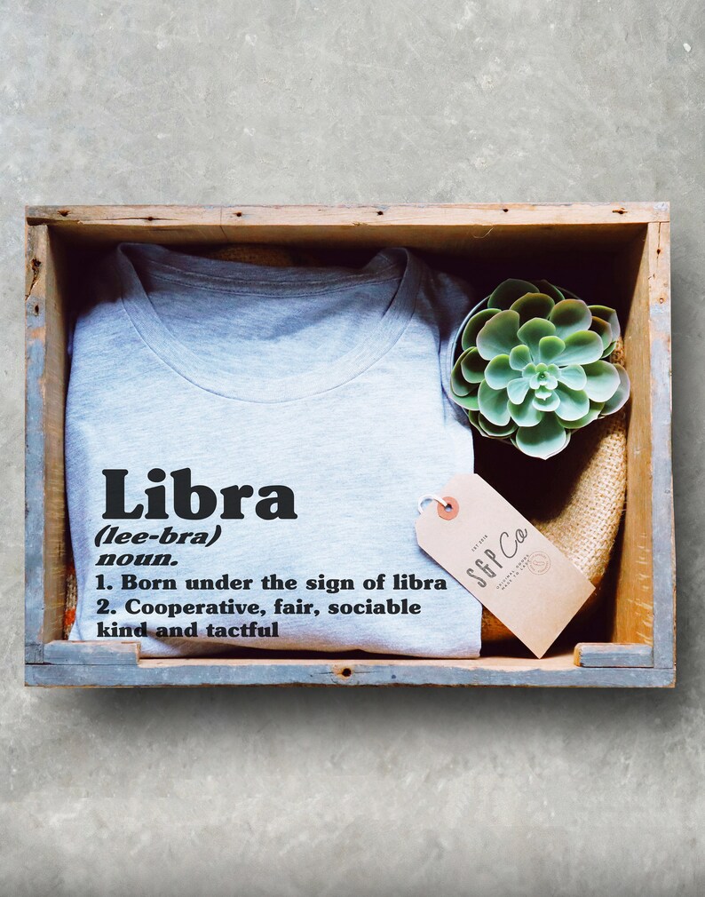 May include: A gray t-shirt with the word "Libra" in black text and a definition of the word. The t-shirt is in a wooden box with a small succulent plant and a brown tag with the text "SGP Co."