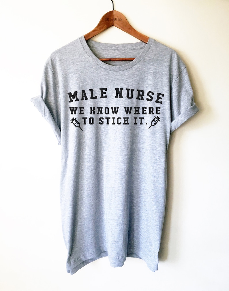 Puede incluir: Camiseta gris con texto negro que dice "Male Nurse We Know Where To Stick It." Dos jeringas negras est&aacute;n a cada lado del texto.