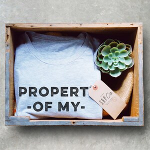 May include: A gray t-shirt with the text "PROPERTY OF MY" in black lettering. The t-shirt is in a wooden crate with a small succulent plant and a brown tag with the text "SGP Co."