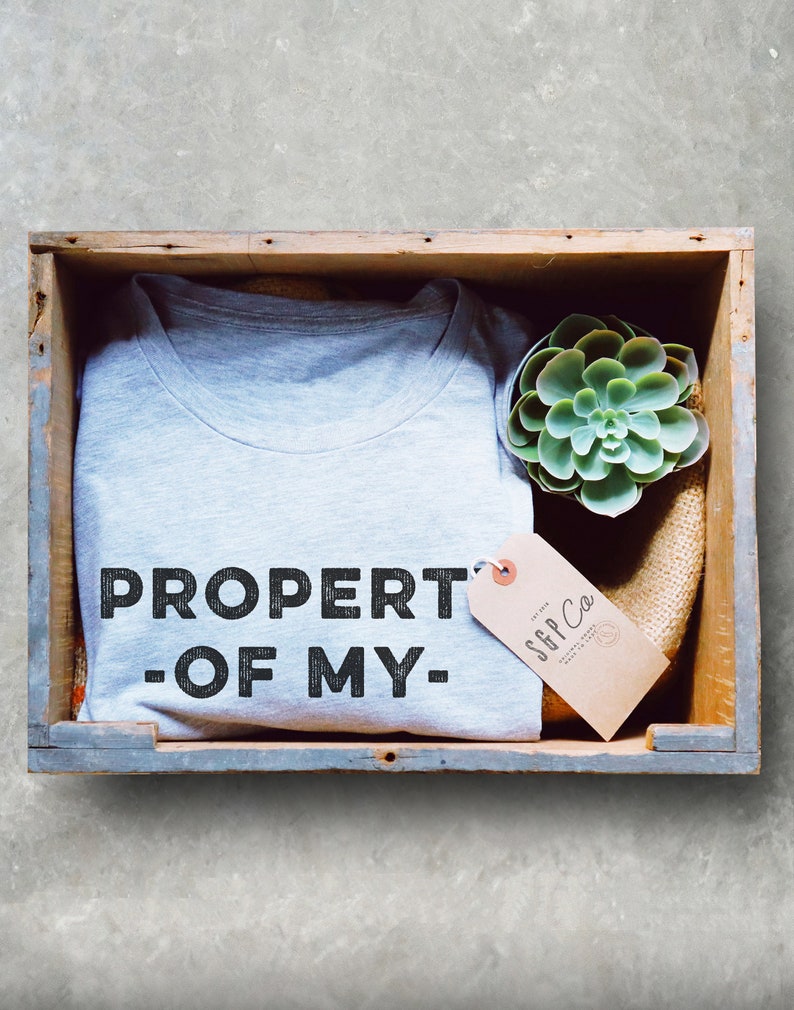 May include: A gray t-shirt with the text "PROPERTY OF MY" in black lettering. The t-shirt is in a wooden crate with a small succulent plant and a brown tag with the text "SGP Co."