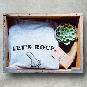May include: A gray t-shirt with the text "LET'S ROCK" and a drawing of a knee bone. The shirt is in a wooden crate with a small green succulent plant and a brown tag that says "SGP Co."