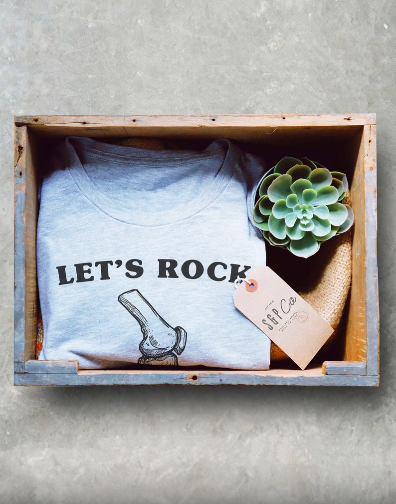 May include: A gray t-shirt with the text "LET'S ROCK" and a drawing of a knee bone. The shirt is in a wooden crate with a small green succulent plant and a brown tag that says "SGP Co."