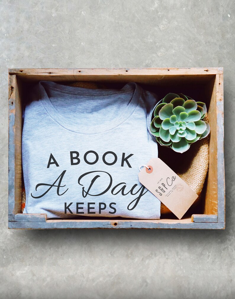 May include: A gray t-shirt with the text "A BOOK A DAY KEEPS" printed on it. The t-shirt is in a wooden crate with a small succulent plant and a brown tag that says "SFF Co."