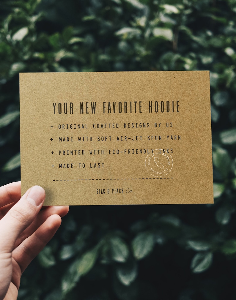 May include: A brown card with the text "YOUR NEW FAVORITE HOODIE" and a list of features: "ORIGINAL CRAFTED DESIGNS BY US", "MADE WITH SOFT AIR-JET SPUN YARN", "PRINTED WITH ECO-FRIENDLY INKS", "MADE TO LAST". The card also has a small round stamp with the text "STAG & PEACH Co."