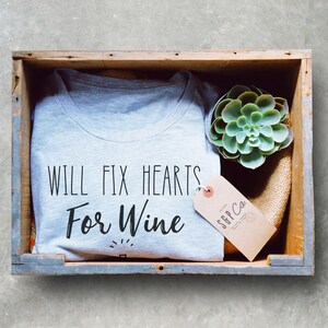 May include: A gray t-shirt with the text "Will Fix Hearts For Wine" in black lettering. The shirt is in a wooden crate with a small succulent plant and a brown tag with the text "S&P Co."