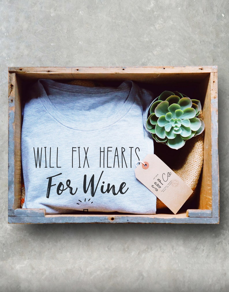 May include: A gray t-shirt with the text "Will Fix Hearts For Wine" in black lettering. The shirt is in a wooden crate with a small succulent plant and a brown tag with the text "S&P Co."