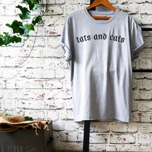 May include: A grey t-shirt with the text "tats and cats" printed in black gothic font.