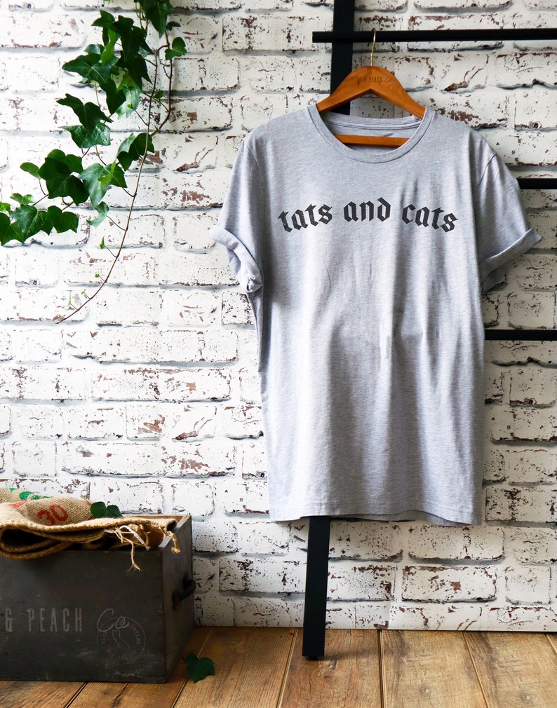 May include: A grey t-shirt with the text "tats and cats" printed in black gothic font.