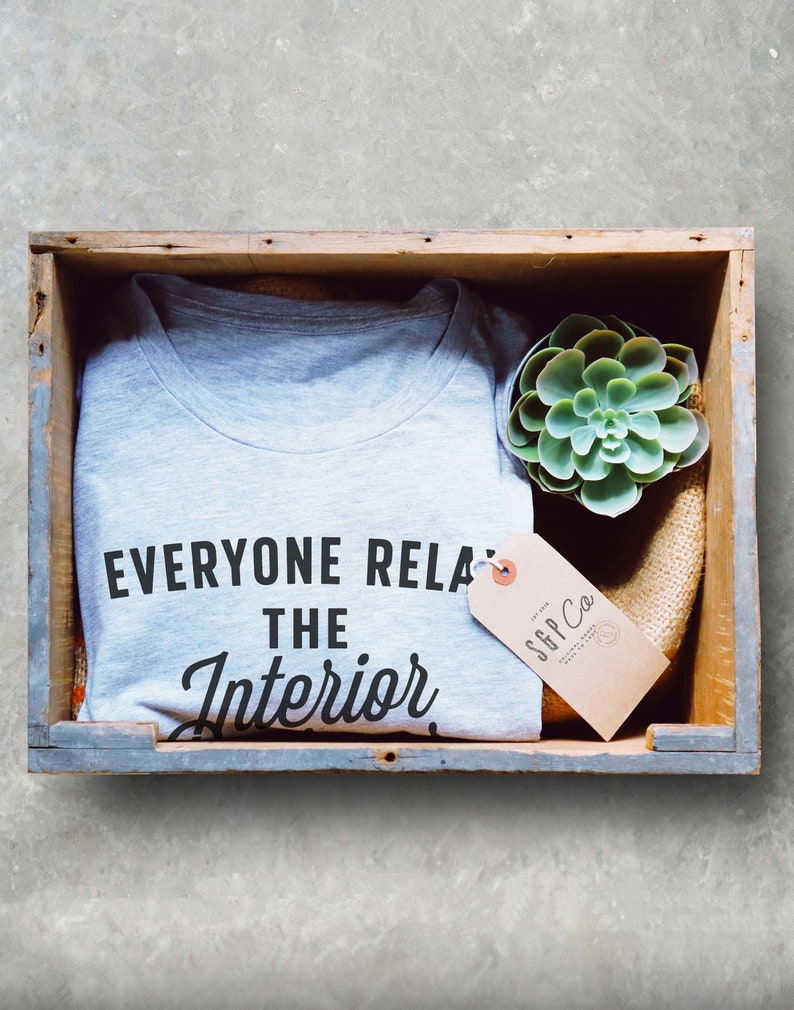 Op de afbeelding: Een grijs T-shirt met de tekst "Everyone Relax The Interior" in zwarte letters. Het T-shirt zit in een houten kist met een kleine vetplant en een bruin label met de tekst "SGP Co."