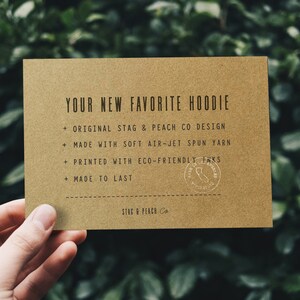 Puede incluir: Una tarjeta marr&oacute;n con el texto "YOUR NEW FAVORITE HOODIE" y una lista de caracter&iacute;sticas: "ORIGINAL STAG & PEACH CO DESIGN", "MADE WITH SOFT AIR-JET SPUN YARN", "PRINTED WITH ECO-FRIENDLY INKS", "MADE TO LAST". La tarjeta tambi&eacute;n tiene un sello que dice "MADE IN LOS ANGELES". En la parte inferior de la tarjeta, dice "STAG & PEACH Co."