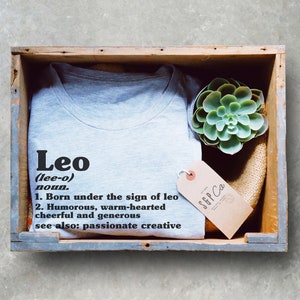 May include: A gray t-shirt with the word "Leo" and a definition of the name. The shirt is in a wooden box with a succulent plant and a brown tag that says "SGP Co."