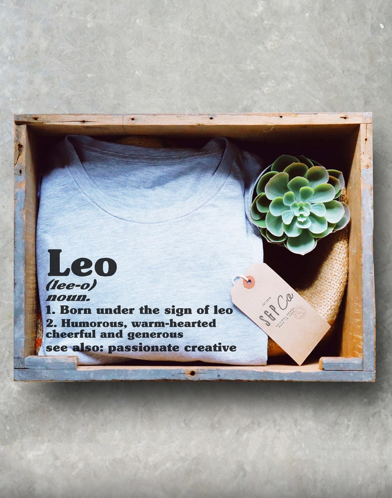 May include: A gray t-shirt with the word "Leo" and a definition of the name. The shirt is in a wooden box with a succulent plant and a brown tag that says "SGP Co."