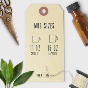 May include: A tan tag displays "MUG SIZES" with illustrations of mugs and the text "11 OZ CAPACITY" and "15 OZ CAPACITY". The tag is surrounded by scissors, a small brown bottle, green leaves, and a spool of thread.