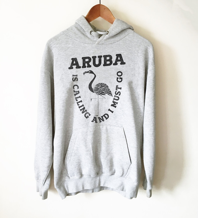 Pu&ograve; includere: Felpa con cappuccio grigia con una stampa grafica nera di un fenicottero e il testo "Aruba is calling and I must go."