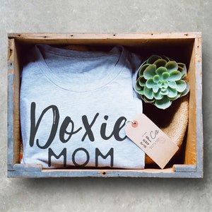 May include: A gray t-shirt with the text "Doxie Mom" in black lettering. The shirt is in a wooden crate with a small succulent plant and a brown tag with the text "SGP Co."