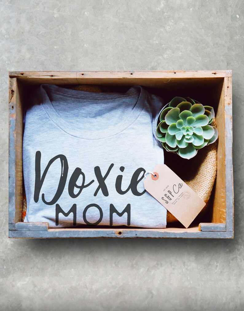 May include: A gray t-shirt with the text "Doxie Mom" in black lettering. The shirt is in a wooden crate with a small succulent plant and a brown tag with the text "SGP Co."