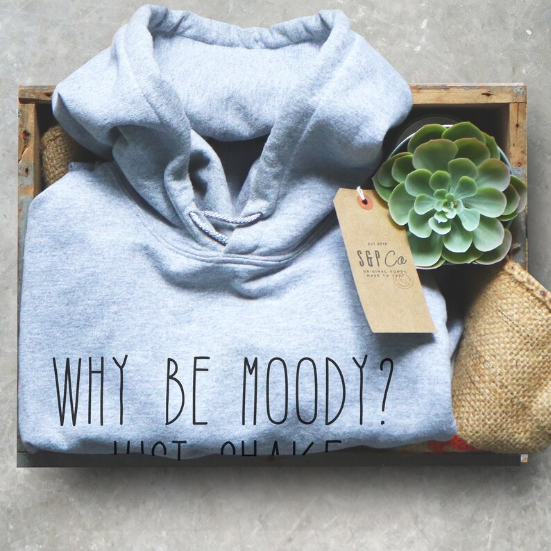 May include: A light gray hoodie with the text "WHY BE MOODY? JUST SHAKE" printed on the front. The hoodie is in a wooden crate with a small succulent plant and a brown tag with the text "SGP Co. ORIGINAL GOODS MADE IN ITALY" on it.