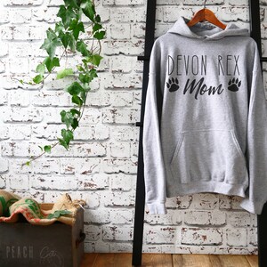May include: A gray hoodie with the text "DEVON REX Mom" and paw prints. The hoodie is hanging on a wooden hanger on a black ladder against a white brick wall. Green ivy hangs from the top left.