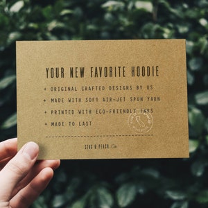 Op de afbeelding: Een bruine kaart met de tekst "YOUR NEW FAVORITE HOODIE" en een lijst met kenmerken: "ORIGINAL CRAFTED DESIGNS BY US", "MADE WITH SOFT AIR-JET SPUN YARN", "PRINTED WITH ECO-FRIENDLY INKS", "MADE TO LAST". De kaart heeft ook een stempel met de tekst "MADE IN LOS ANGELES". Onderaan de kaart staat "STAG & PEACH Co."