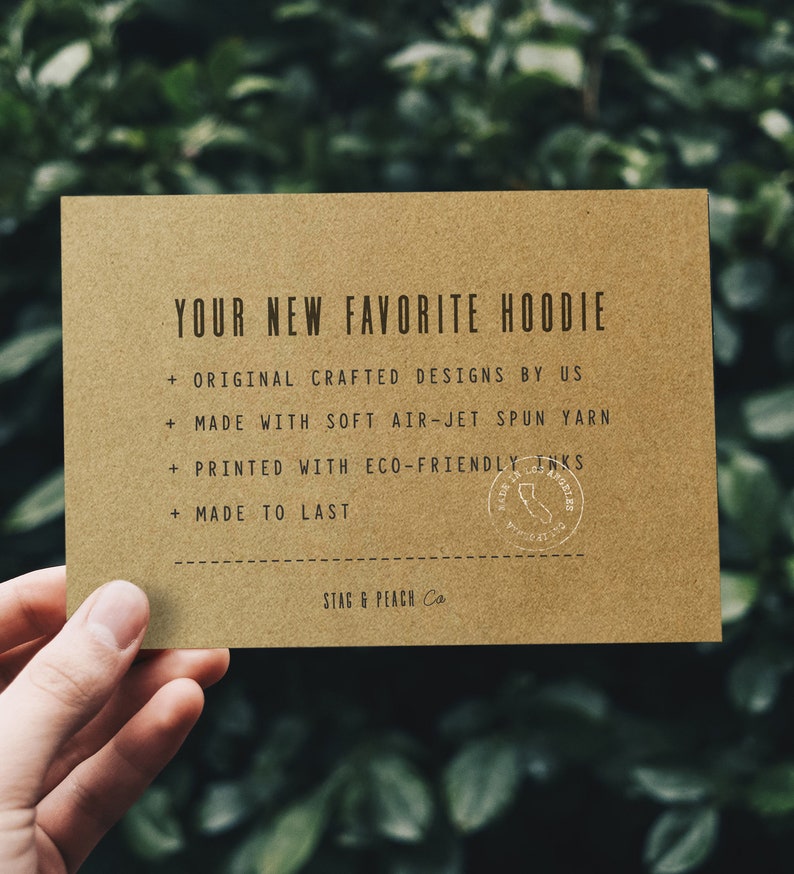 Op de afbeelding: Een bruine kaart met de tekst "YOUR NEW FAVORITE HOODIE" en een lijst met kenmerken: "ORIGINAL CRAFTED DESIGNS BY US", "MADE WITH SOFT AIR-JET SPUN YARN", "PRINTED WITH ECO-FRIENDLY INKS", "MADE TO LAST". De kaart heeft ook een stempel met de tekst "MADE IN LOS ANGELES". Onderaan de kaart staat "STAG & PEACH Co."