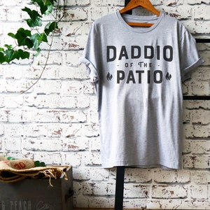 May include: A gray t-shirt with the text "Daddio of the Patio" printed on it. The text is in black and has a flame design on either side of the word "Patio".