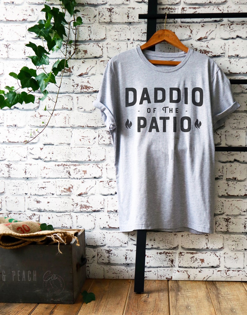 May include: A gray t-shirt with the text "Daddio of the Patio" printed on it. The text is in black and has a flame design on either side of the word "Patio".