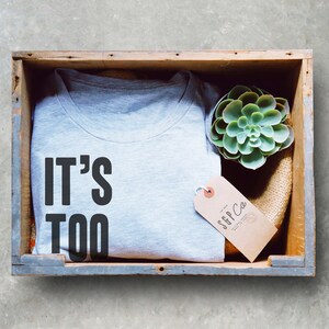 Puede incluir: Una camiseta gris con el texto "IT'S TOO" impreso en ella. La camiseta est&aacute; doblada y colocada en una caja de madera con una peque&ntilde;a planta suculenta y una etiqueta marr&oacute;n con el texto "SFP Co."