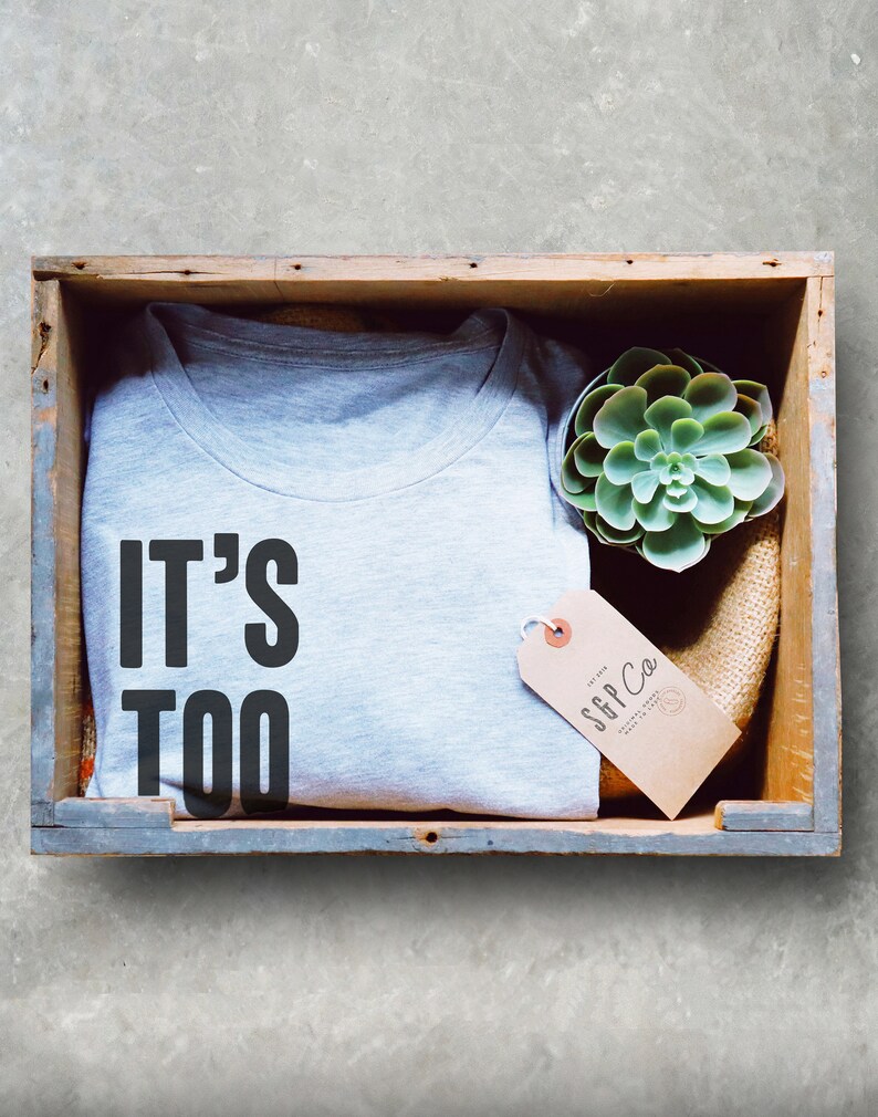 Puede incluir: Una camiseta gris con el texto "IT'S TOO" impreso en ella. La camiseta est&aacute; doblada y colocada en una caja de madera con una peque&ntilde;a planta suculenta y una etiqueta marr&oacute;n con el texto "SFP Co."