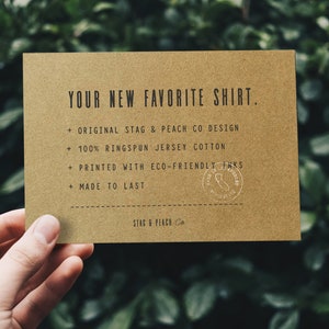 Puede incluir: Una tarjeta marr&oacute;n con el texto "YOUR NEW FAVORITE SHIRT." y una lista de caracter&iacute;sticas: "ORIGINAL STAG & PEACH CO DESIGN", "100% RINGSPUN JERSEY COTTON", "PRINTED WITH ECO-FRIENDLY INKS", "MADE TO LAST". Un sello blanco con el texto "STAG & PEACH CO LOS ANGELES" se encuentra en la esquina inferior derecha.