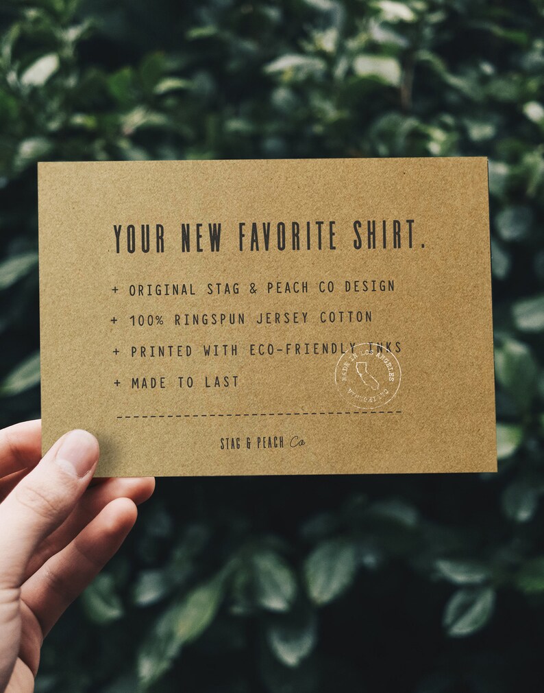 Puede incluir: Una tarjeta marr&oacute;n con el texto "YOUR NEW FAVORITE SHIRT." y una lista de caracter&iacute;sticas: "ORIGINAL STAG & PEACH CO DESIGN", "100% RINGSPUN JERSEY COTTON", "PRINTED WITH ECO-FRIENDLY INKS", "MADE TO LAST". Un sello blanco con el texto "STAG & PEACH CO LOS ANGELES" se encuentra en la esquina inferior derecha.