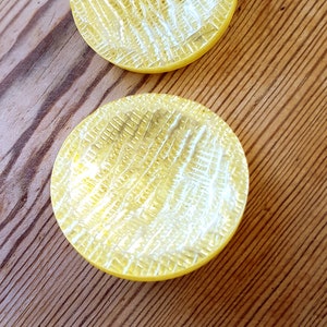 Puede incluir: Dos posavasos de vidrio amarillo con una superficie texturizada. Los posavasos son redondos y tienen un borde ligeramente elevado.