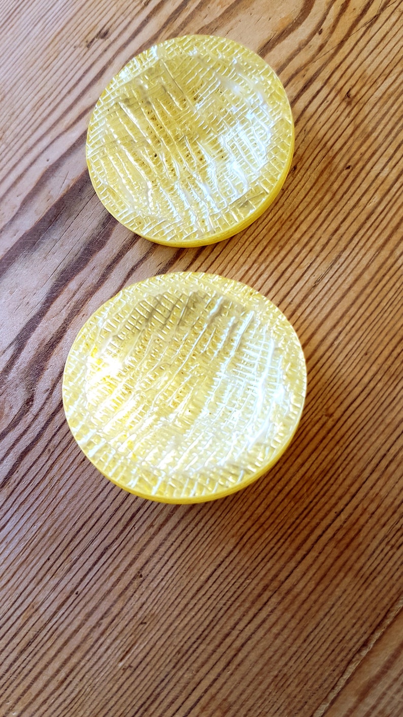 Puede incluir: Dos posavasos de vidrio amarillo con una superficie texturizada. Los posavasos son redondos y tienen un borde ligeramente elevado.