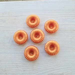 May include: Six orange fabric buttons with a raised center and a circular design. The buttons are arranged in a semi-circle on a light wood surface.