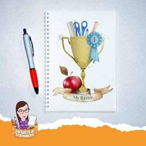 May include: A white spiral-bound notebook featuring a gold trophy design, school supplies, and a red apple. The notebook has the text "Mr Baxter" on a banner. A silver and red pen is to the left.
