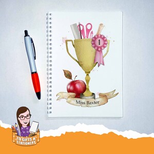 May include: A white spiral-bound notebook featuring a gold trophy design, scissors, and an apple. A red and silver pen lies beside the notebook. The notebook has the text "Miss Baxter" on a banner.