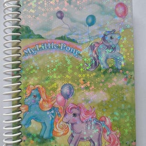 May include: A spiral bound notebook with a holographic cover featuring three My Little Pony characters with colourful balloons. The background is a green field with flowers and a blue sky.
