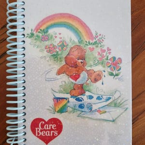 Puede incluir: Una libreta de espiral blanca con un logotipo de Care Bears en la portada. La portada presenta un arcoíris, flores y un Care Bear pintando un cuadro con pintura azul.