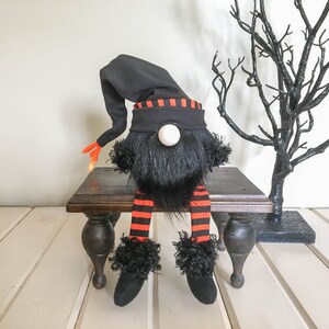 May include: A Halloween gnome doll with a black hat and beard, orange and black striped legs, and black shoes. The gnome is sitting on a small, dark wooden table. A black decorative tree is in the background.
