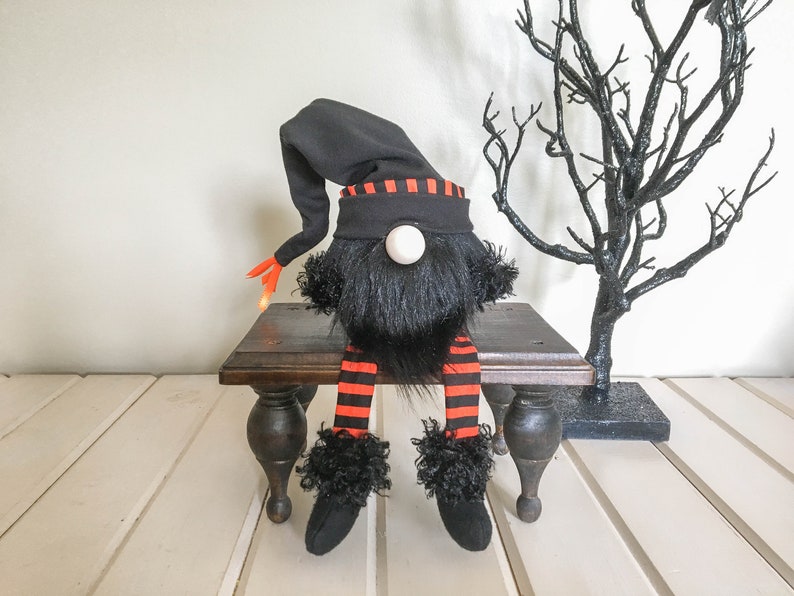 May include: A Halloween gnome doll with a black hat and beard, orange and black striped legs, and black shoes. The gnome is sitting on a small, dark wooden table. A black decorative tree is in the background.