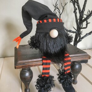 May include: A Halloween gnome doll with a black hat and beard, orange and black striped legs, and a wooden nose. The gnome is sitting on a small, dark wooden bench. Decorative black branches are in the background.