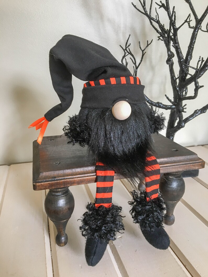 May include: A Halloween gnome doll with a black hat and beard, orange and black striped legs, and a wooden nose. The gnome is sitting on a small, dark wooden bench. Decorative black branches are in the background.