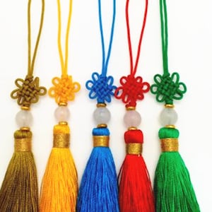 May include: Five colourful tassel keychains with a knotted design. The tassels are green, yellow, blue, red, and green. Each keychain has a small jade-coloured bead and a gold-coloured bead.
