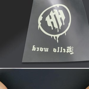 May include: A white vinyl sticker with a dripping circle design and the text "brow village" in a gothic font. The sticker is on a black background and is being peeled off of a sheet of clear plastic. The text "PET with Stickiness" is visible in the background.