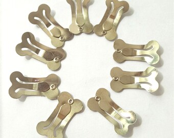 May include: Ten gold-colored metal hair clips shaped like bones. The clips are arranged in a circle on a white background.