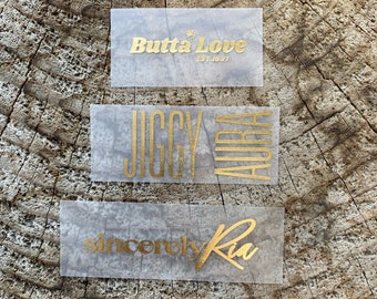 Op de afbeelding: Vier transparante labels met gouden letters. Het bovenste label heeft de tekst "Butta Love Est. 1997". Het tweede label heeft de tekst "Jiggy Aura". Het derde label heeft de tekst "sincerely Ra". Het onderste label heeft de tekst "NO PRIVACY STUDIOS".