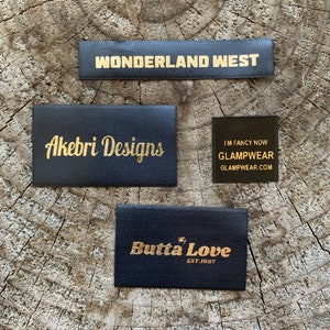 Könnte beinhalten: Vier schwarze Stoffetiketten mit goldenem Text auf einer Holzoberfläche angeordnet. Die Etiketten lauten "WONDERLAND WEST", "Akebri Designs", "I'M FANCY NOW GLAMPWEAR.COM" und "Butta Love EST.1997".