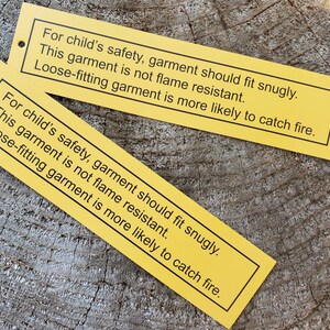 May include: Two yellow warning labels with black text. The text reads: "For child's safety, garment should fit snugly. This garment is not flame resistant. Loose-fitting garment is more likely to catch fire."