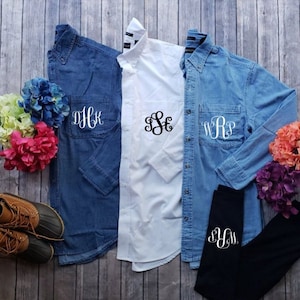 May include: Three denim shirts with different monograms embroidered on the pockets. The shirts are laid out on a wooden surface. One shirt is white and the other two are blue.