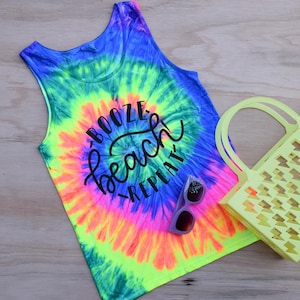 May include: A tie-dye tank top with the text "Booze Beach Repeat" in black lettering. The tank top is a bright neon green, blue, orange, and pink. A pair of sunglasses and a yellow plastic tote bag are also in the image.